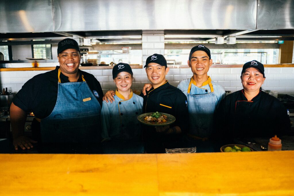 A group of diverse chefs smiling and posing together in a modern restaurant kitchen.